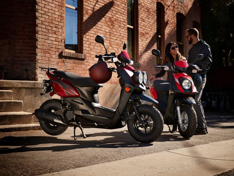 Two double seater scooters parked in front of a brick building. Two people are standing behind one of the scooters.