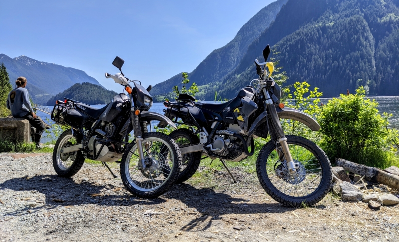 A DR 650 and DRZ parked along side the ocean on a gravel Road
