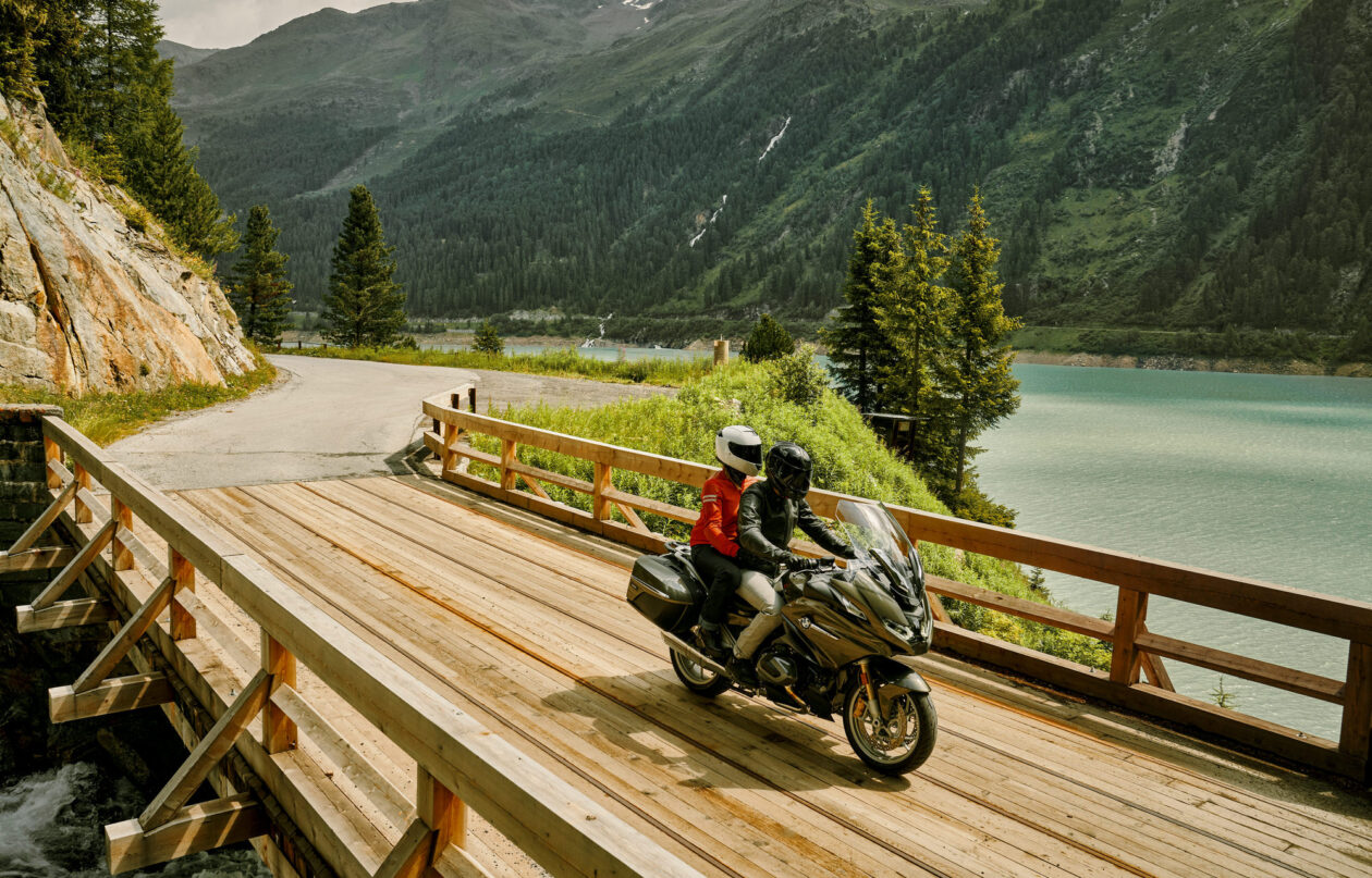 A couple riding a BMW R 1250 RT TFT over a wooden bridge in the mountains.